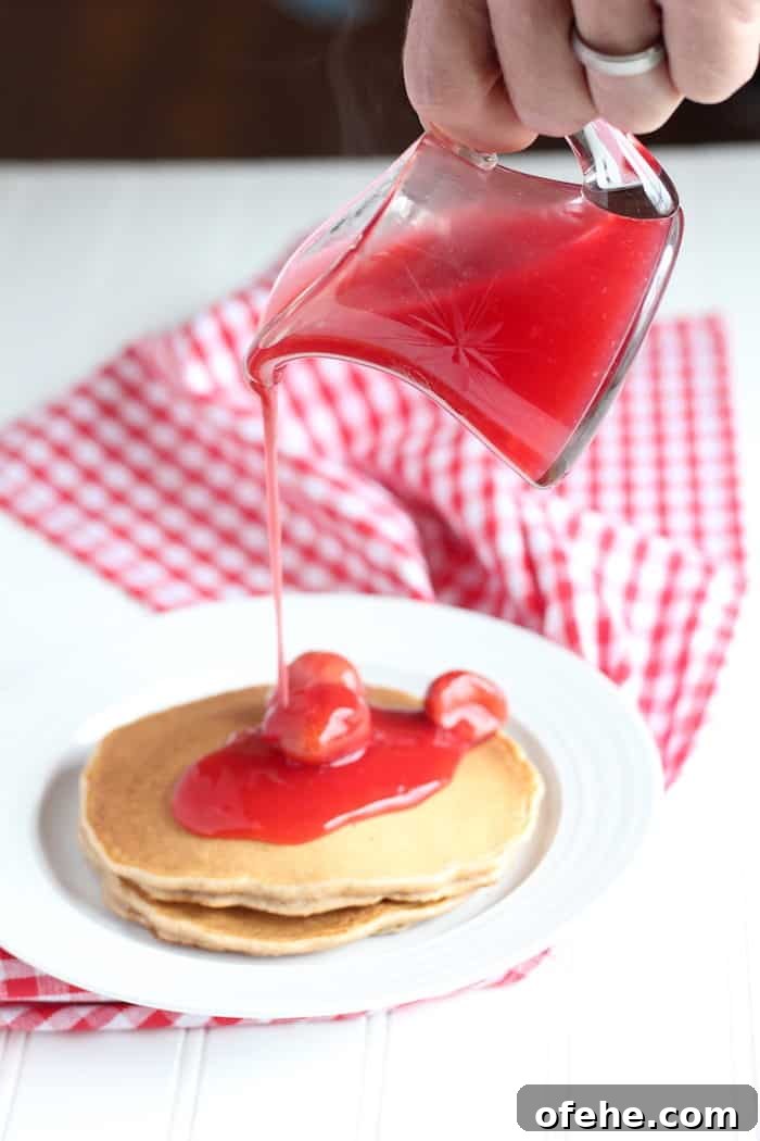 Homemade Strawberry Syrup Recipe for Pancakes & Drinks 4 Image shows a hand pouring strawberry syrup over a plate of pancakes