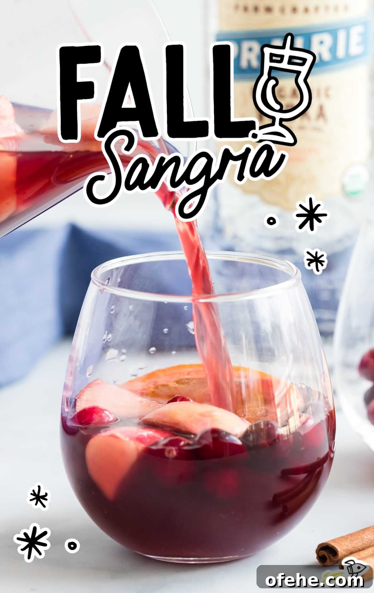 Front view of a glass of fall sangria mid-pour with fresh fruit and cinnamon sticks, vodka bottle in background