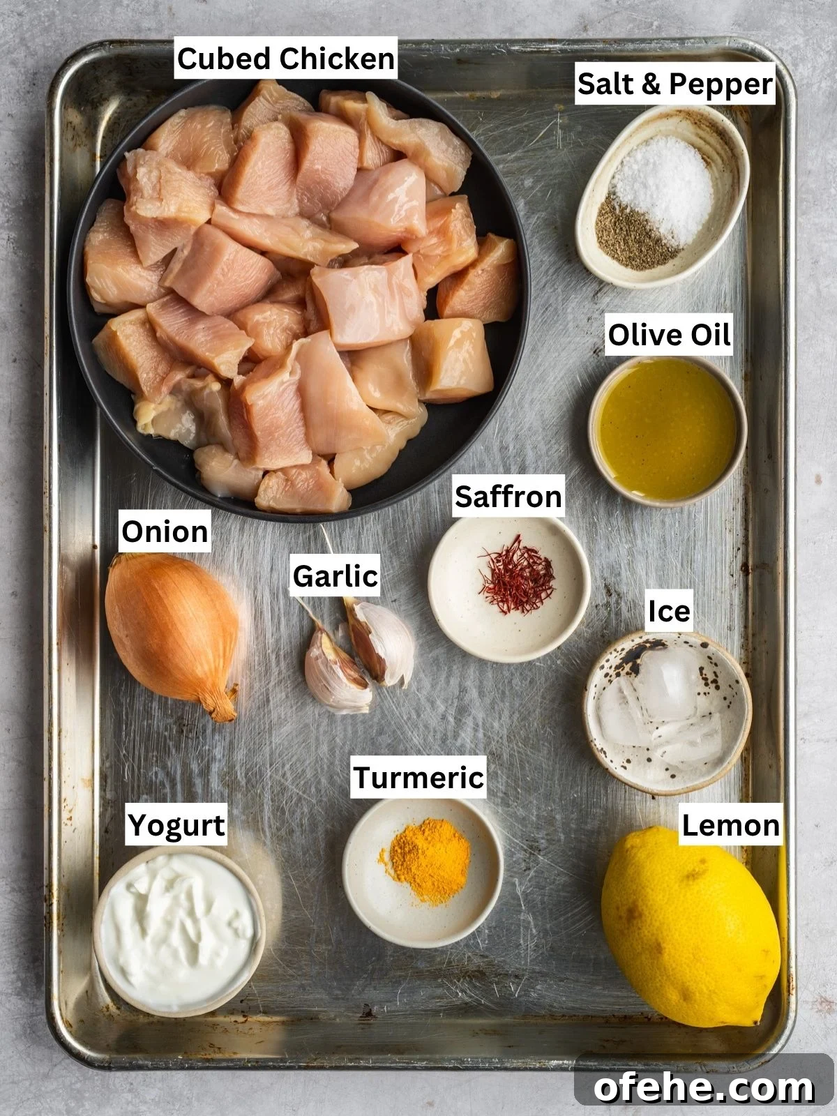 Persian Saffron Joojeh Kabob Recipe — Grilled Chicken Kabab 3 A metal tray with labeled ingredients for Joojeh Kabob: a bowl of cubed chicken, salt and pepper, olive oil, an onion, garlic cloves, saffron threads, yogurt, ground turmeric, a lemon, and ice cubes.