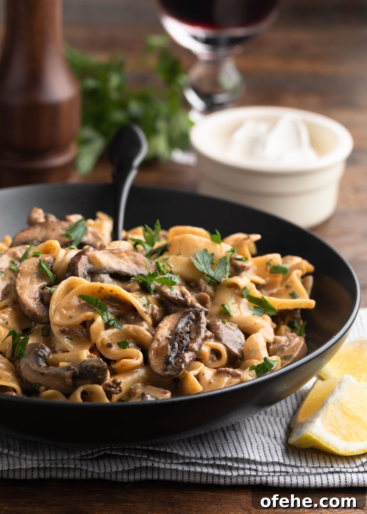 One-Pan Creamy Ground Beef Stroganoff Recipe 2 ground beef stroganoff in a black ceramic bowl with a black fork, in front of a bowl of sour cream, a glass of red wine, and a pepper mill