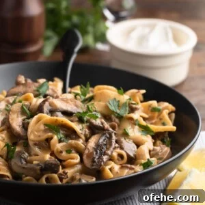 One-Pan Creamy Ground Beef Stroganoff Recipe 11 ground beef stroganoff in a black ceramic bowl with a black fork, in front of a bowl of sour cream, a glass of red wine, and a pepper mill