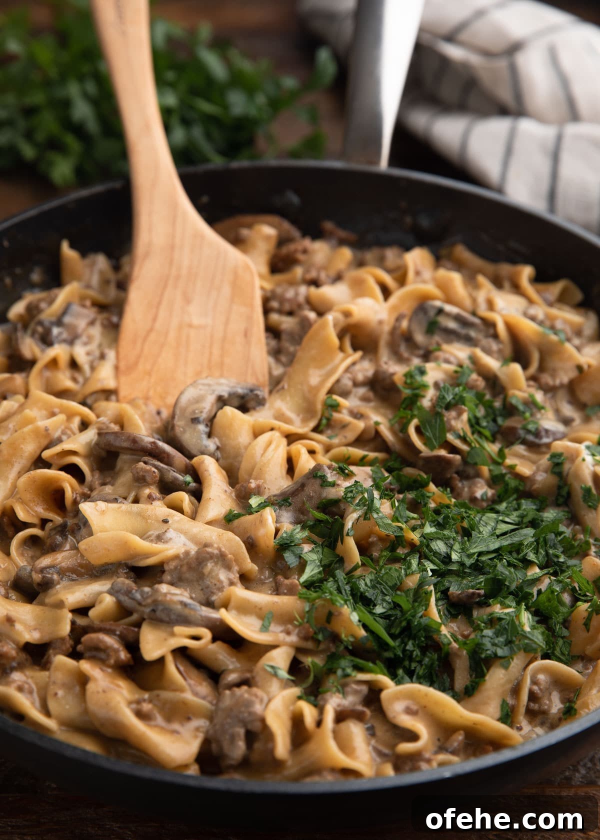 One-Pan Creamy Ground Beef Stroganoff Recipe 8 Skillet filled with creamy ground beef stroganoff, topped with a generous handful of chopped parsley and stirred with a wooden spoon.