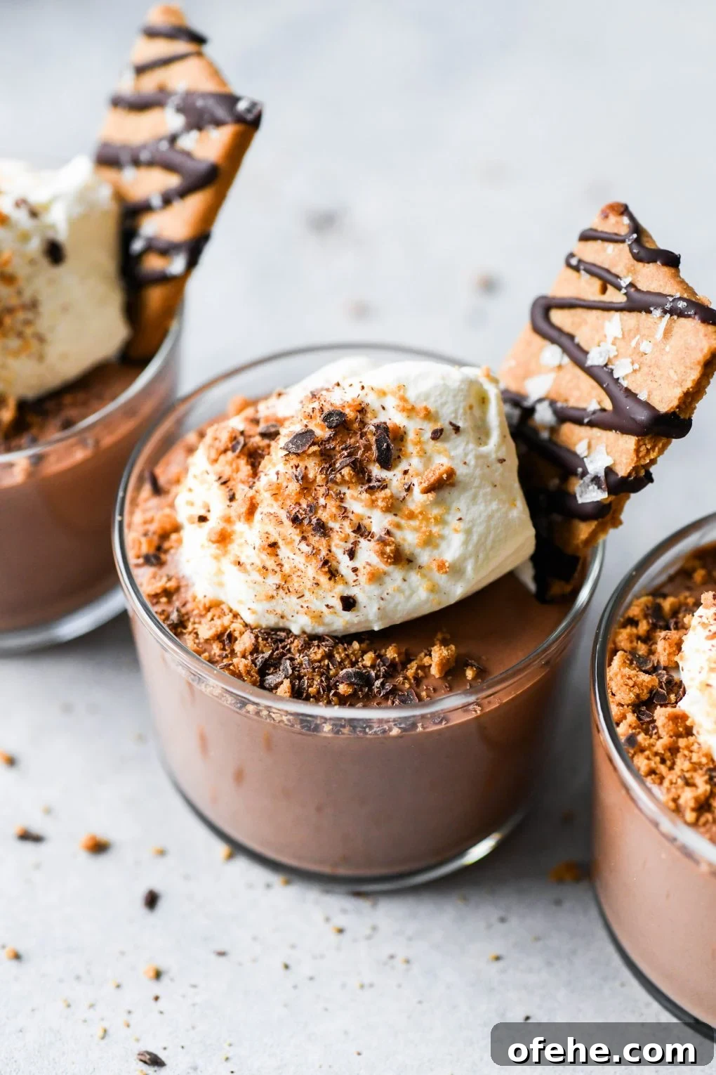 Overhead shot of three vegan mousse cups in an angled row on a light colored background topped with whipped cream and chocolate drizzled graham crackers. 