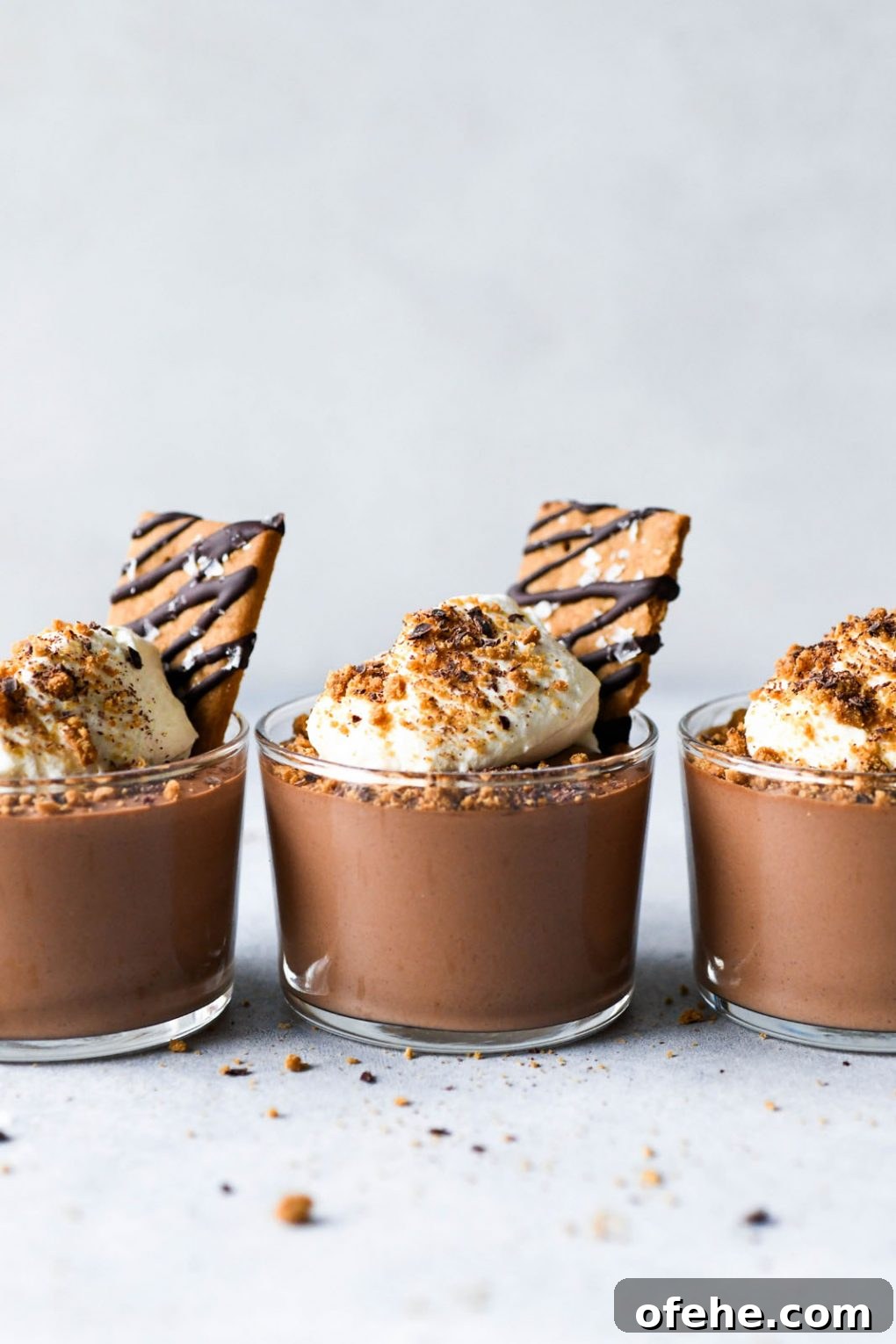 Straight on shot of three vegan mousse cups in a row on a light colored background topped with whipped cream and chocolate drizzled graham crackers. 