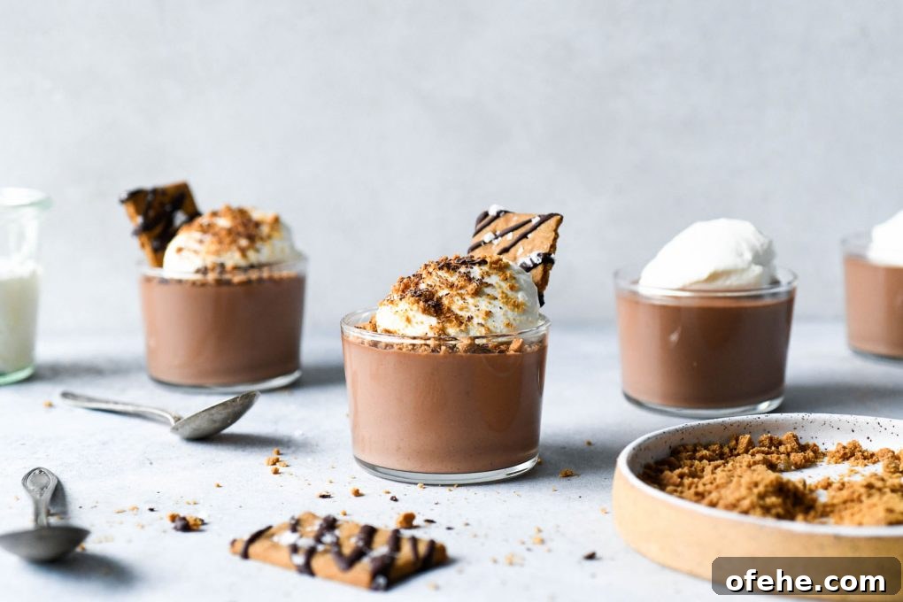 A straight on shot of a several staggered chocolate mousse cups topped with whipped cream, graham cracker crumbs, and a chocolate drizzled graham cracker topped with flaky sea salt. On a light colored background surrounded be several silver spoons, and a small speckled plate with crumbled graham crackers.