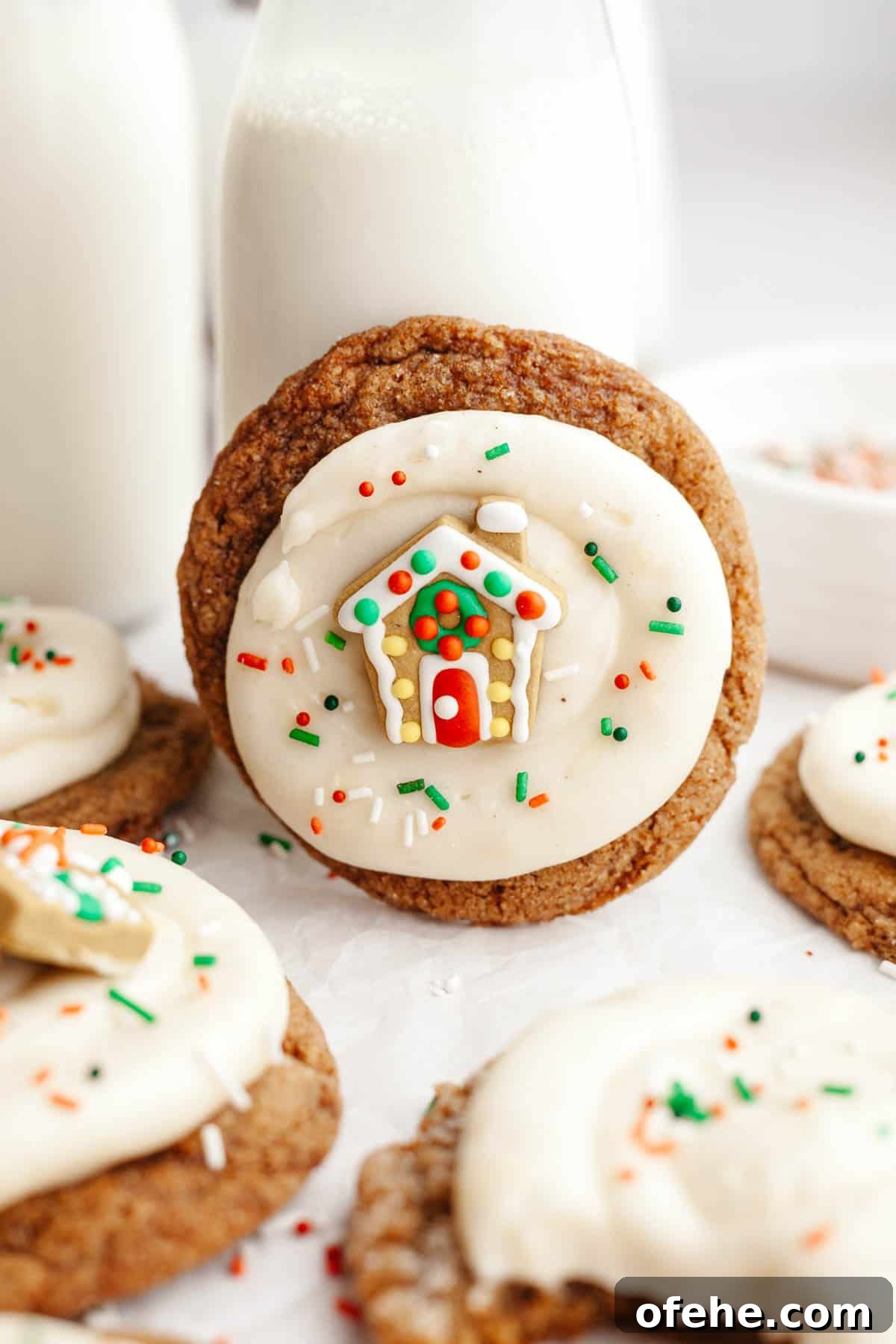 Soft Frosted Gingerbread Cookies with Cream Cheese Frosting Recipe 12 A close up of gingerbread cookies frosted with cream cheese frosting and a small gingerbread house decoration.