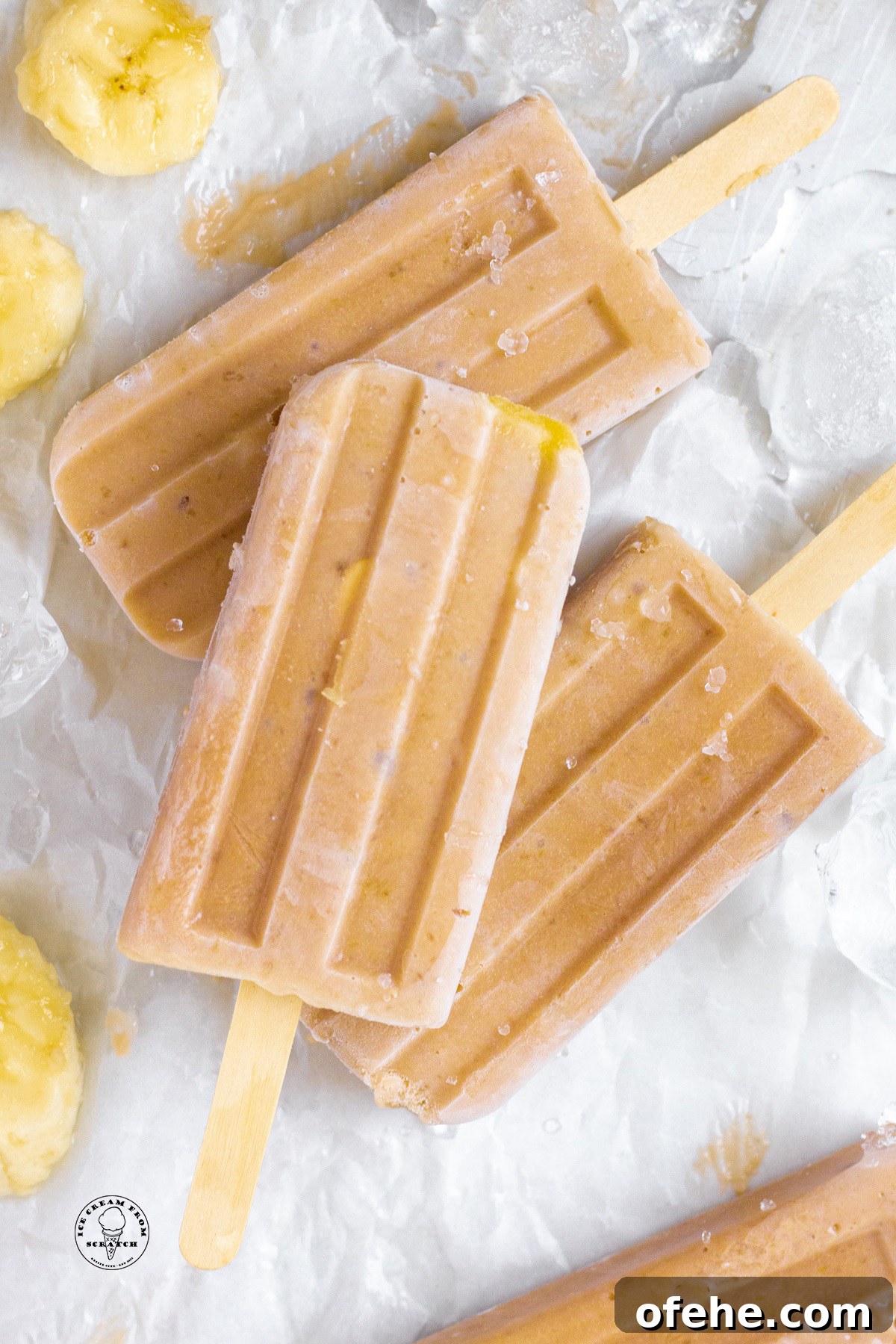 Homemade Banana Popsicles Recipe — Creamy Frozen Treats 5 Three banana popsicles on a piece of parchment paper with slices of ripe banana
