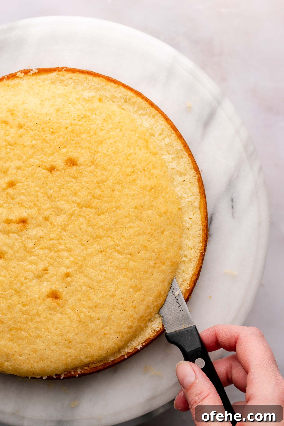 Raspberry Coconut Cake Recipe with Creamy Coconut Frosting 12 A hand uses a serrated knife to remove the domed top from a baked cake round.