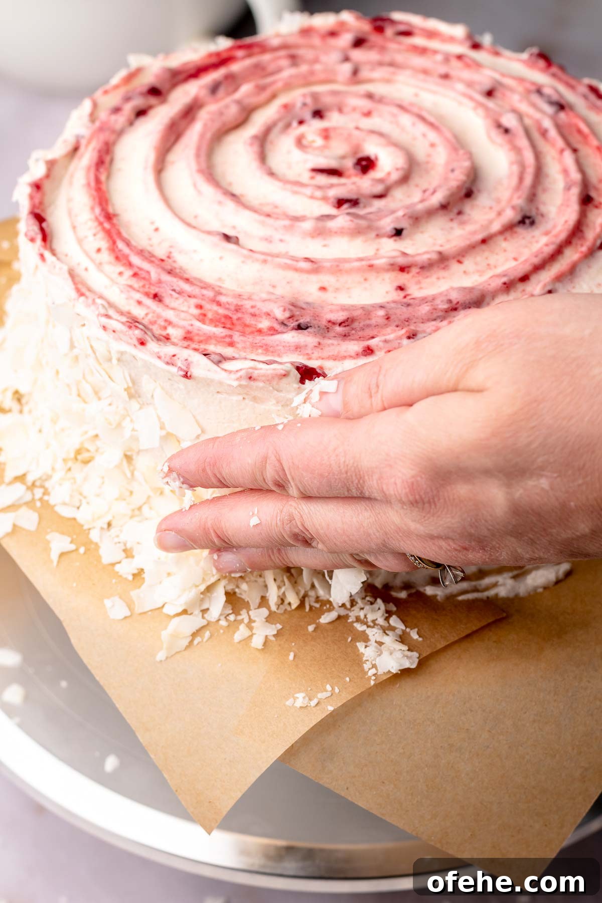 Raspberry Coconut Cake Recipe with Creamy Coconut Frosting 16 A hand adds coconut flakes to the sides of the cake.