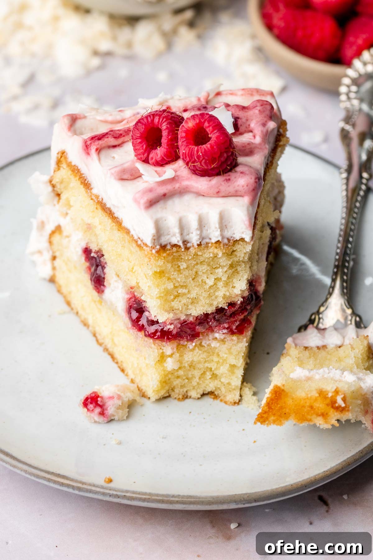 Raspberry Coconut Cake Recipe with Creamy Coconut Frosting 18 A slice of coconut raspberry cake on a plate with a piece on a fork sitting next to it.