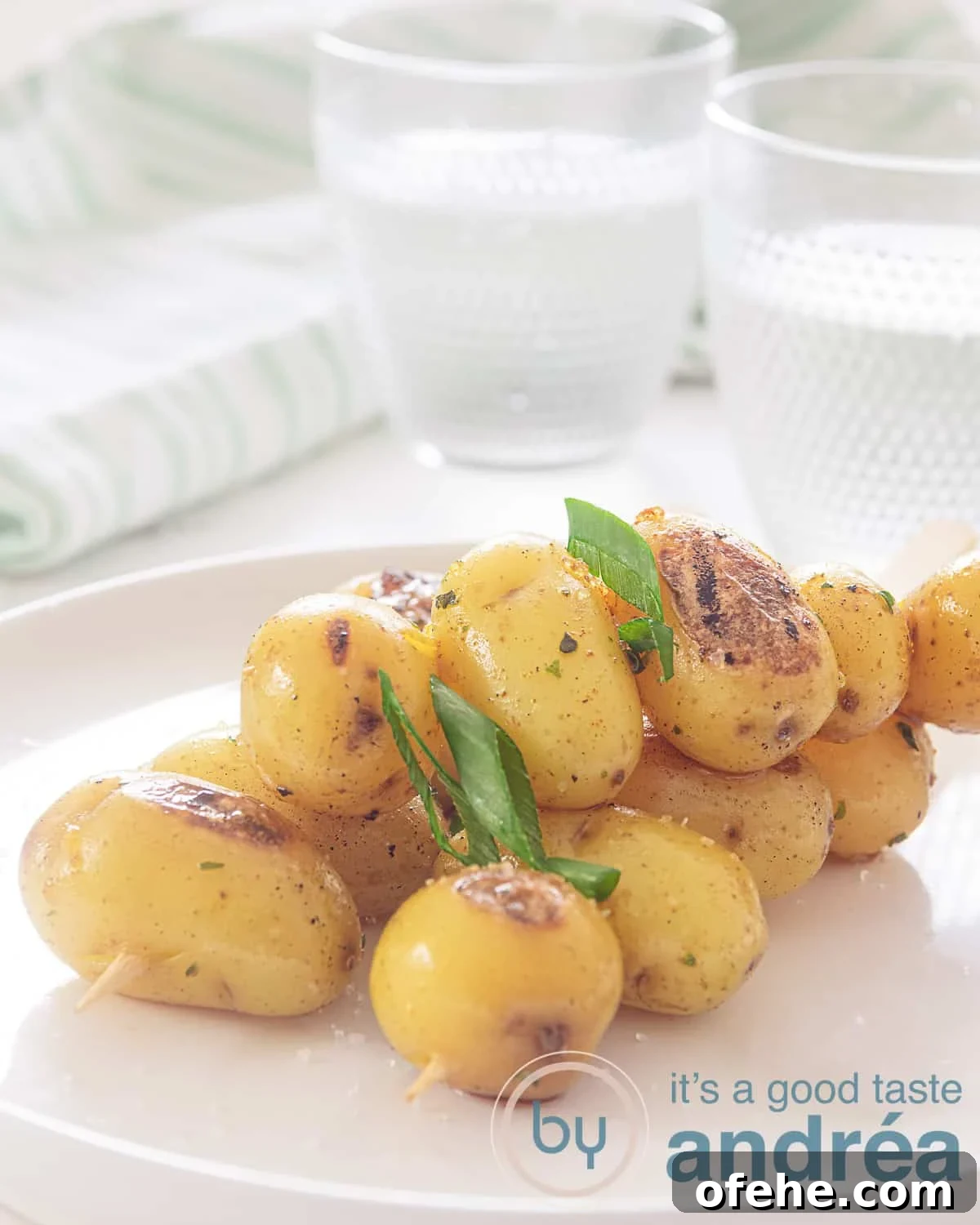 Grilled BBQ Baby Potato Skewers – Savory Summer Side Dish 2 A height photo with a white plate with three skewers with delicious seasoned baby potatoes from the bbq. A white background. In the background two glasses of water and a white and green striped tea towel.