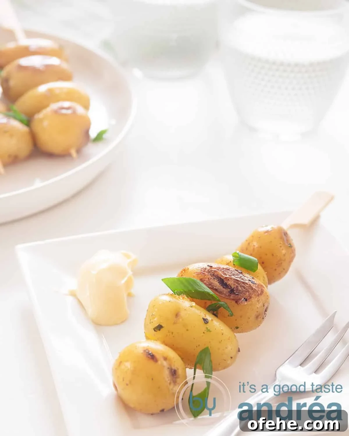 Grilled BBQ Baby Potato Skewers – Savory Summer Side Dish 3 A baby kebab on a white plate with some sauce. In the background more baby potatoes on a stick and a glass of water.