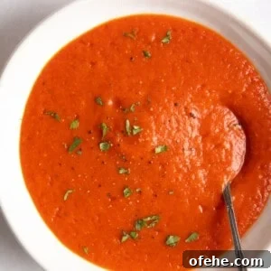 Roasted Red Pepper & Tomato Soup Recipe for Cozy Weeknights 12 red pepper and tomato soup served in a white bowl with a spoon inside.