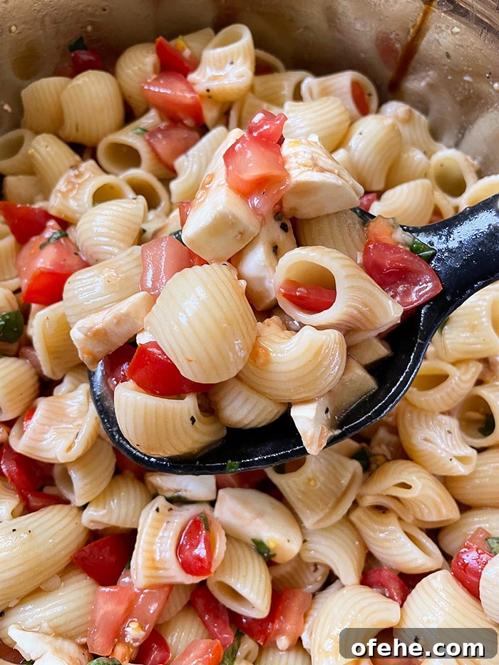 Bruschetta Pasta Salad in a large metal bowl with a black serving spoon held above with a scoop of salad.