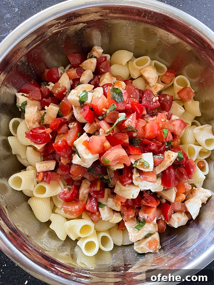 A large metal mixing bowl with small-shaped pasta topped with bruschetta mixed with diced fresh mozzarella.