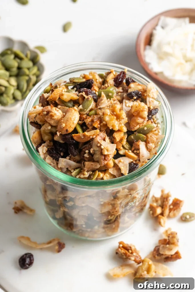 Grain-Free Homemade Granola Recipe with Crunchy Seeds 4 nut and seed grain free granola in a clear french mason jar with two smaller bowls of pepitas and unsweetened coconut flakes on the white table and some of the granola and seeds scattered around.