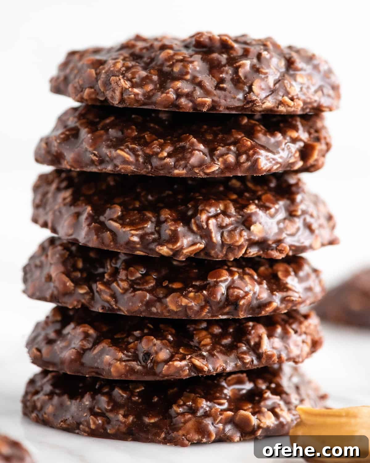 a stack of 6 no bake cookies on a white surface with more cookies and a spoon of peanut butter surrounding it