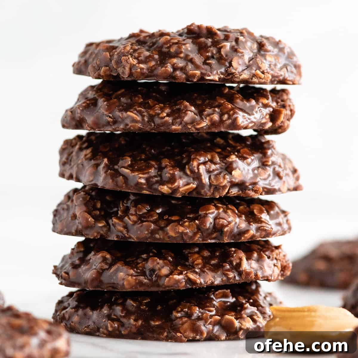 a stack of 6 no bake cookies on a white surface with more cookies and a spoon of peanut butter surrounding it