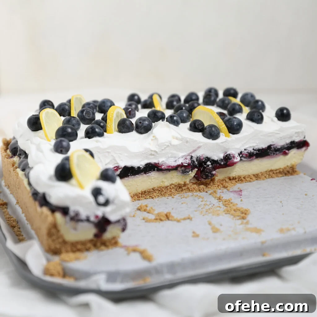 A blueberry lemon delight dessert bar with a graham cracker crust, creamy layer, blueberry topping, and whipped cream. Slices are topped with fresh blueberries and lemon wedges; part of the dessert has been cut and served.