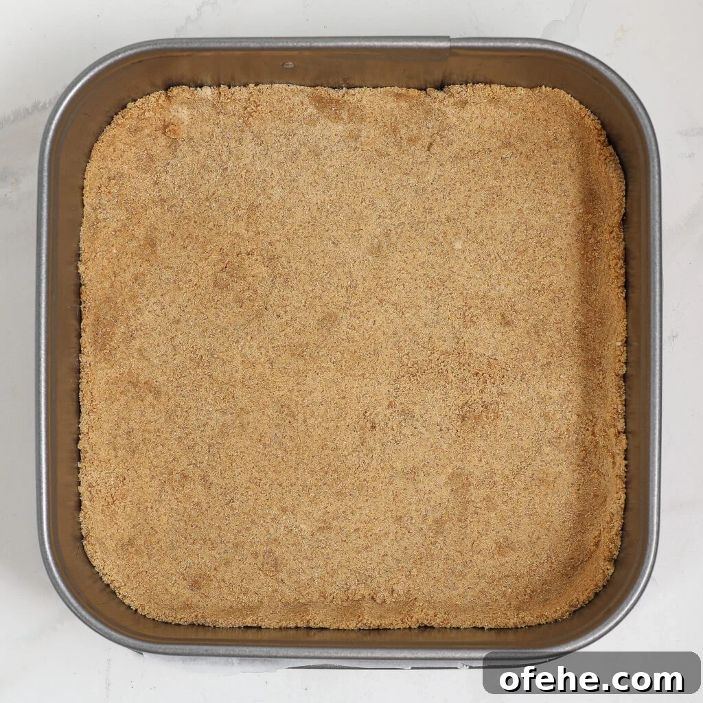 Square baking pan filled with an even layer of light brown, crumbly graham cracker crust mixture, pressed flat against the bottom—perfect for creating a refreshing blueberry lemon delight.