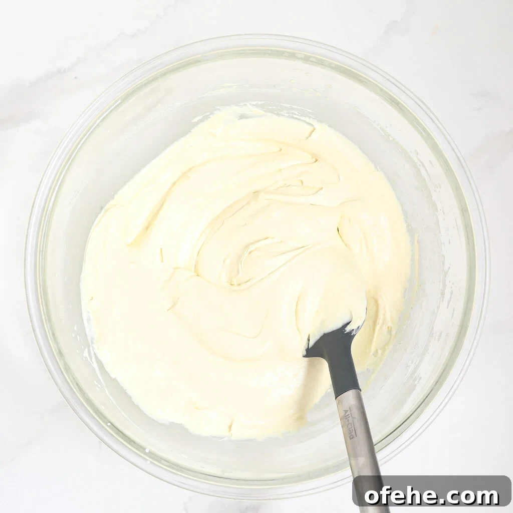A glass bowl filled with creamy, pale batter for a blueberry lemon delight is being mixed with a gray silicone spatula on a white marble surface.