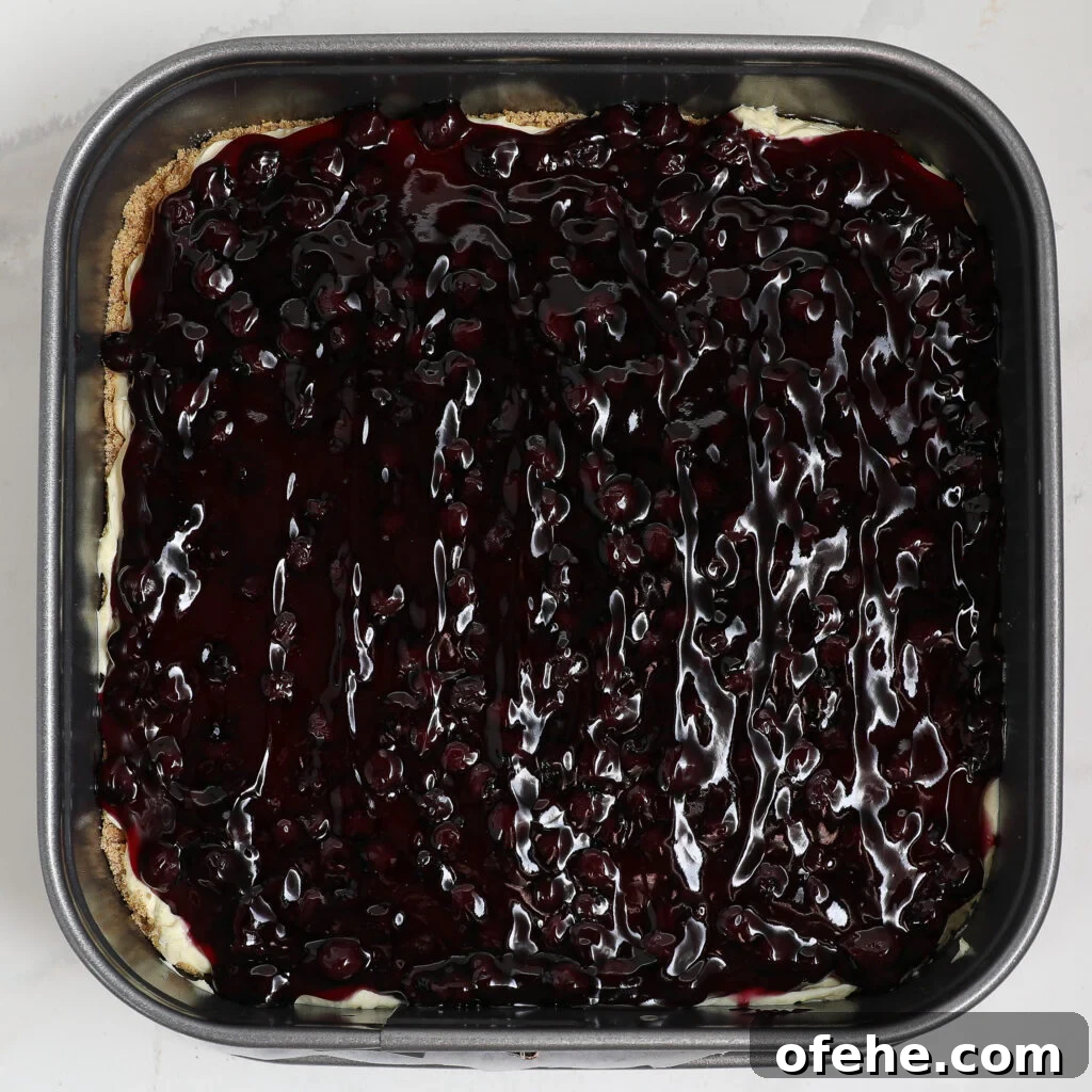 A square baking pan filled with blueberry lemon delight: a dessert topped with glossy, dark purple blueberry sauce over a creamy layer and light crust. The sauce is spread evenly across the surface.