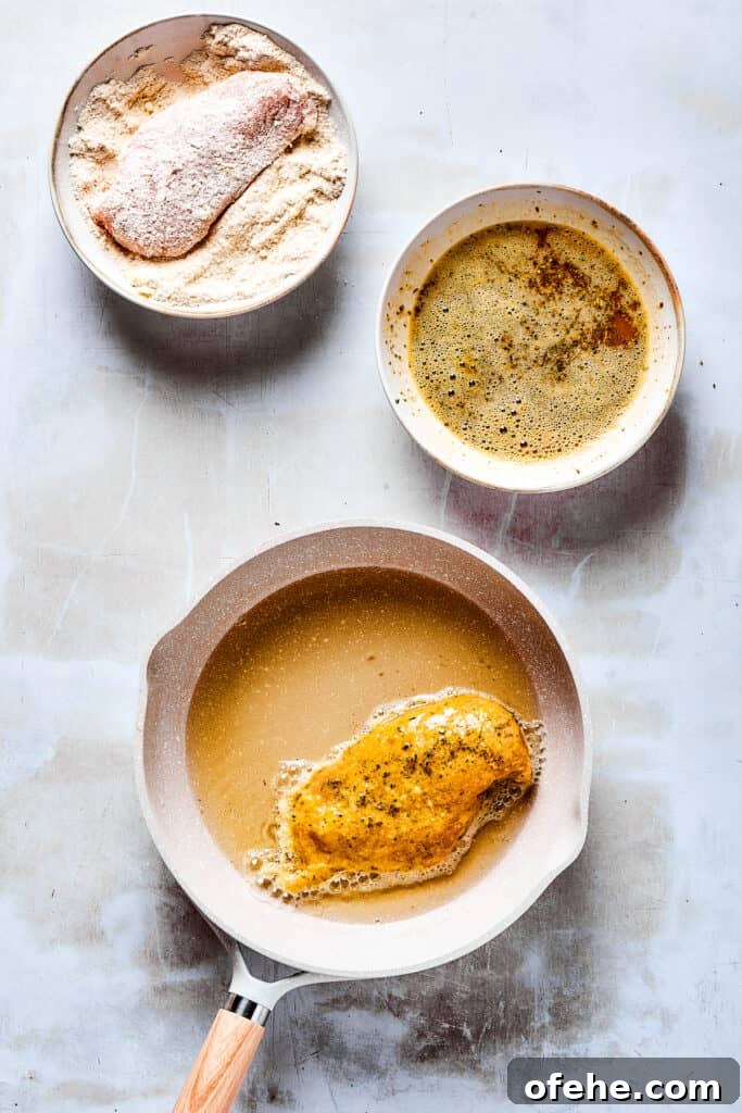 Chicken being breaded and fried. A chicken breast in a dish of dry breading ingredients, next to a dish of wet breading ingredients, and a skillet with one breaded chicken breast cooking in oil.
