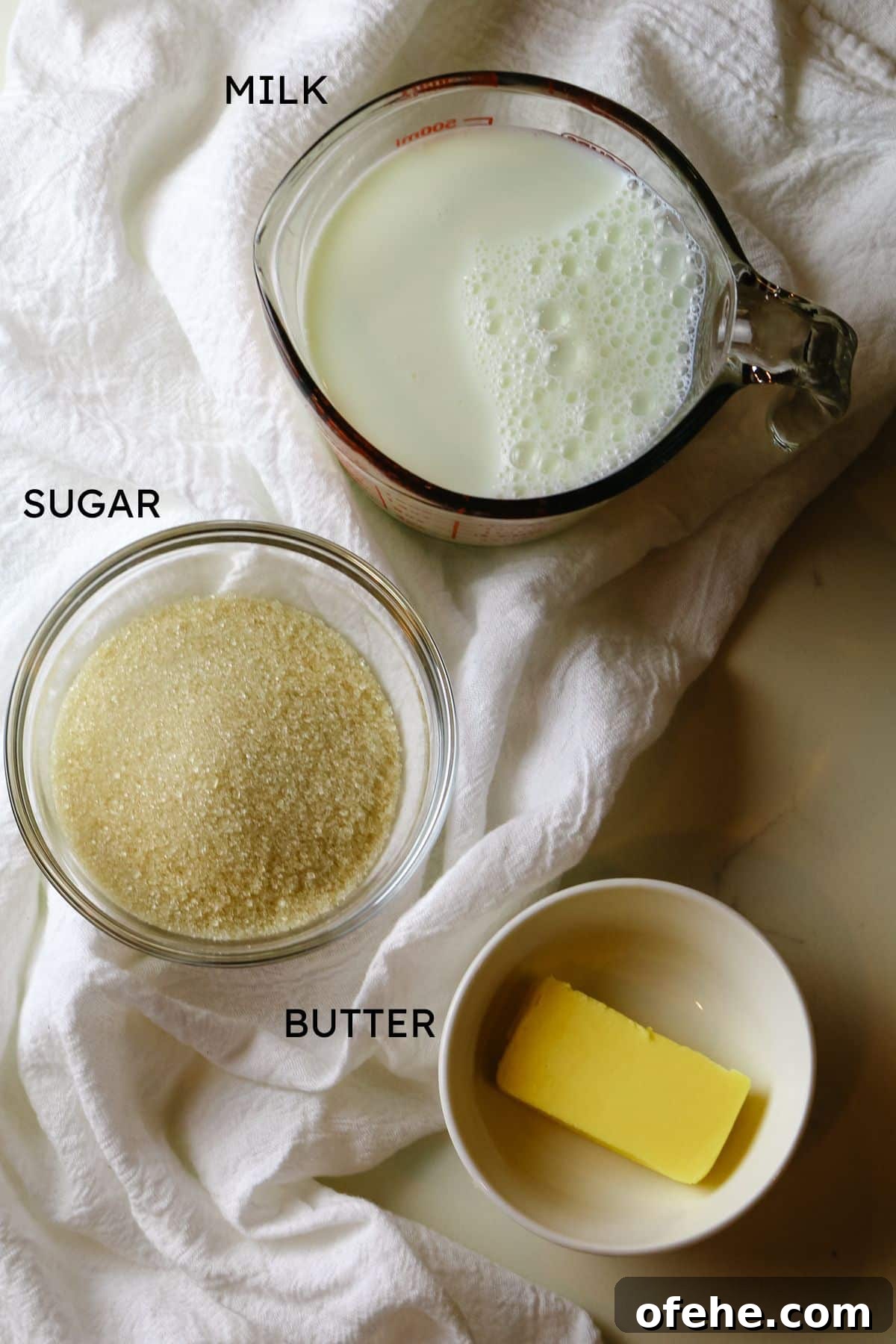an ingredients shot including milk in a measuring cup, sugar is a glass jar, and butter in a bowl.