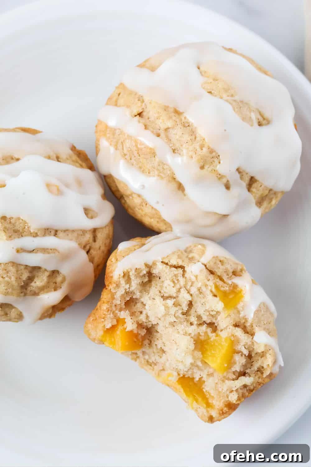 Peach Muffins Made with Bisquick — Quick Summer Treat 4 three easy peach muffins on a white plate, topped with icing. One muffin has a bite taken out of it, showing chunks of yellow peaches in the fluffy interior.