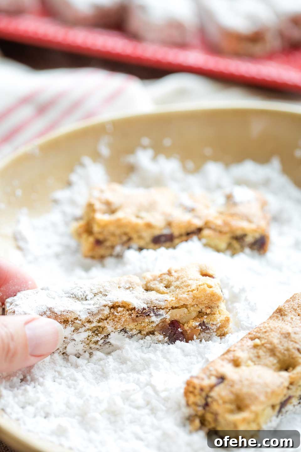 Closeup of three Date Bars in a bowl of powdered sugar, with fingers reaching in to roll one around in it.