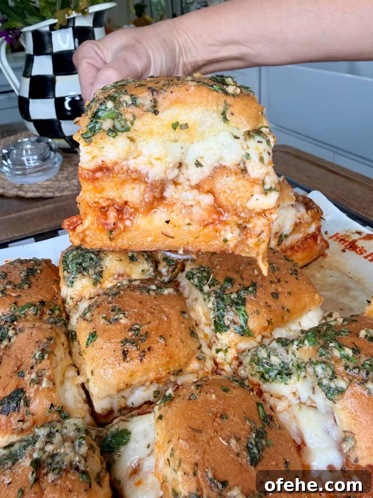 A hand lifts a cheesy, baked sandwich from a tray of pull-apart sliders, topped with herbs and melted cheese. The sandwiches are golden brown and layered with marinara sauce and mozzarella.