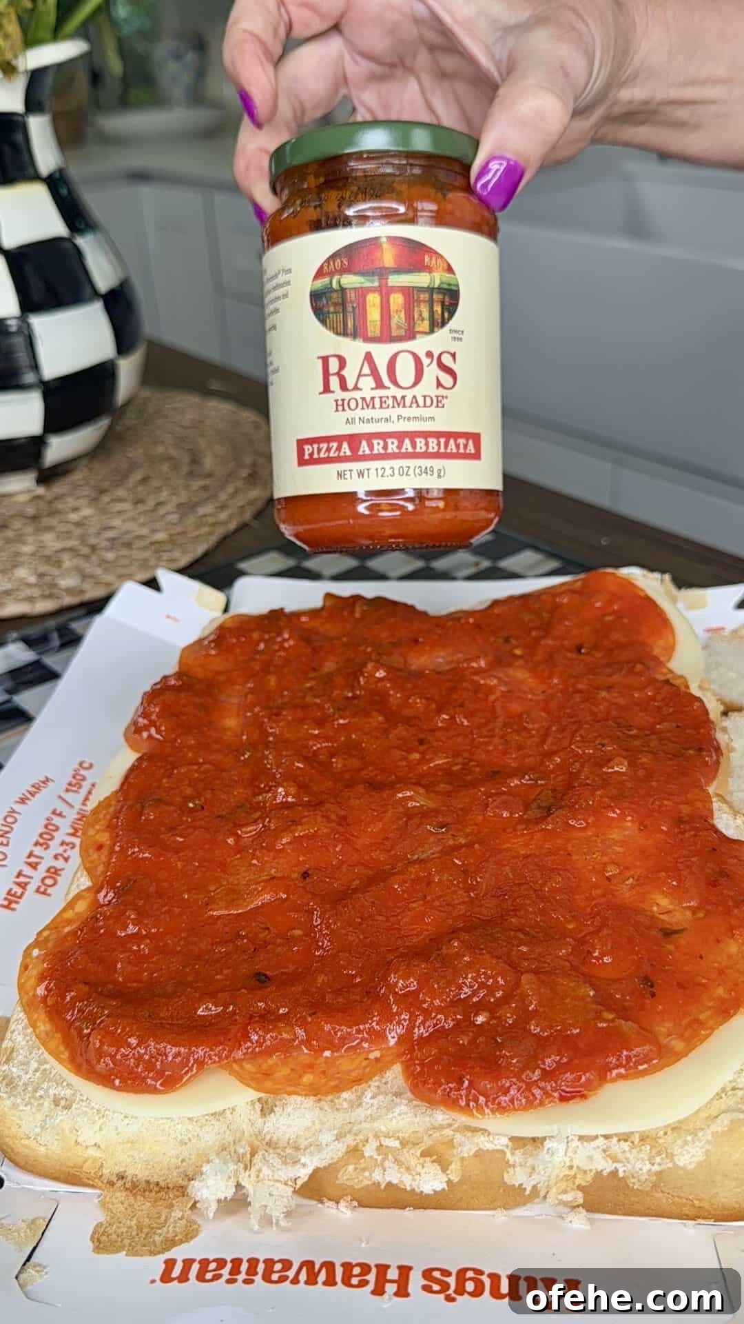 A hand holds a jar of Rao’s Homemade Pizza Arrabbiata sauce above a sandwich bun covered in melted cheese and topped with pizza sauce, on a King’s Hawaiian tray in a kitchen.