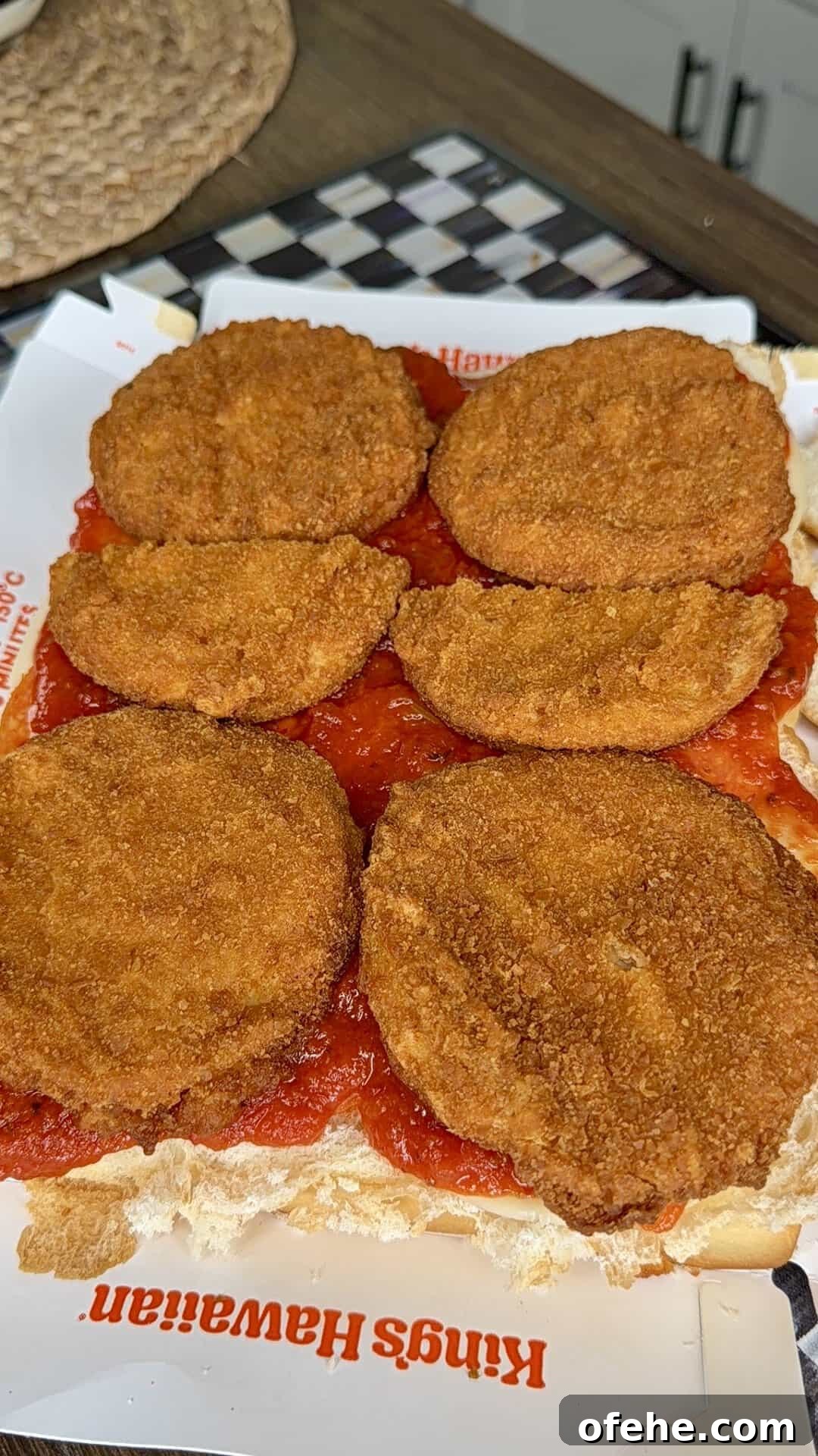 Six breaded and fried eggplant slices are arranged on tomato sauce over a sliced King’s Hawaiian sandwich roll, with a checkered board and woven mat visible in the background.