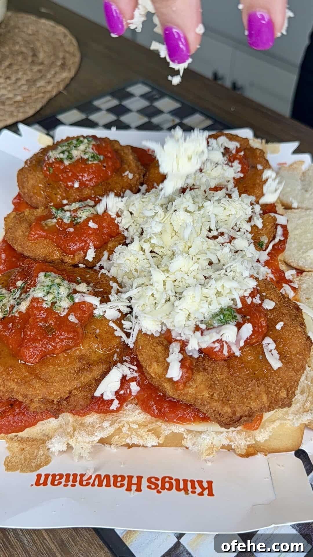 A hand with bright purple nails sprinkles shredded cheese onto a sandwich filled with breaded and fried cutlets topped with marinara sauce, all placed on a hoagie roll on a Kings Hawaiian wrapper.