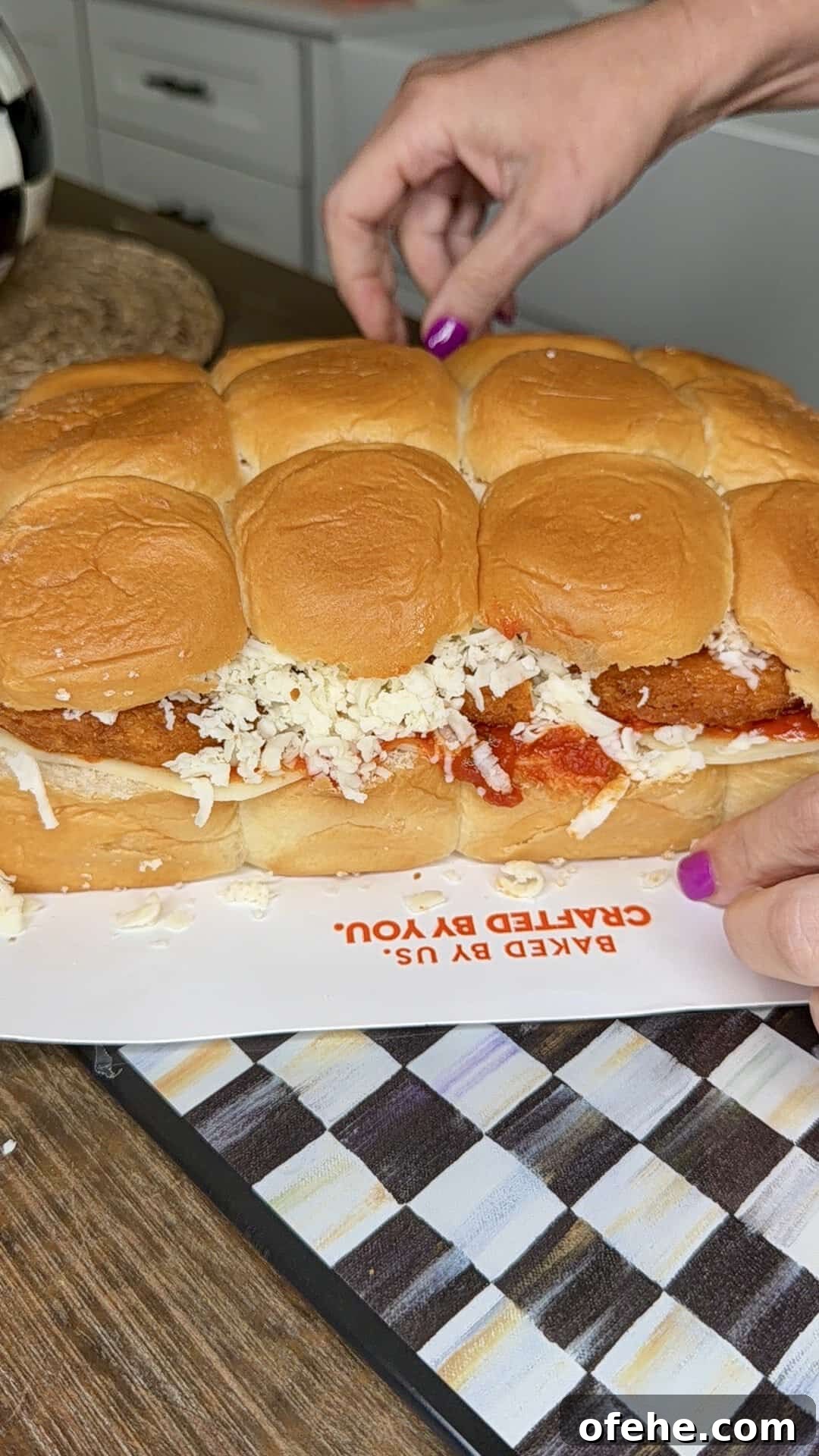 A person with purple nail polish slices a sandwich made of bread rolls filled with breaded chicken, marinara sauce, and shredded cheese on a kitchen counter.