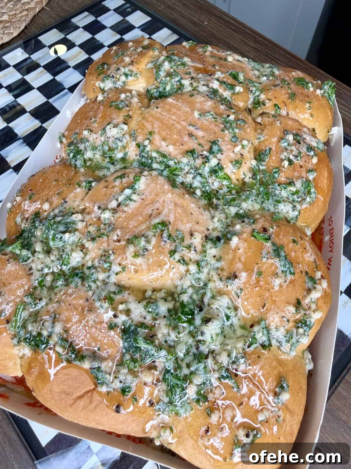A tray of fluffy golden-brown bread rolls topped with melted butter, chopped herbs, and minced garlic, sitting on a checkered black-and-white countertop.