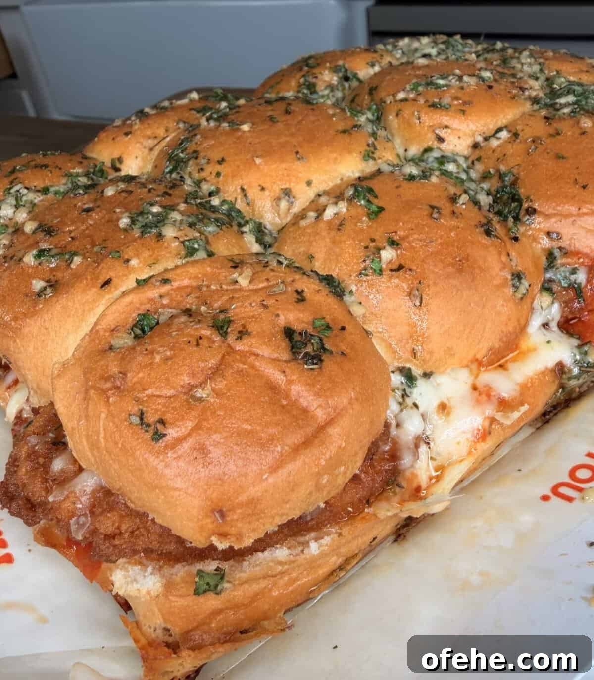 A close-up of a tray of cheesy chicken sliders on soft, golden buns topped with herbs and garlic, with melted cheese and sauce visible between the layers.