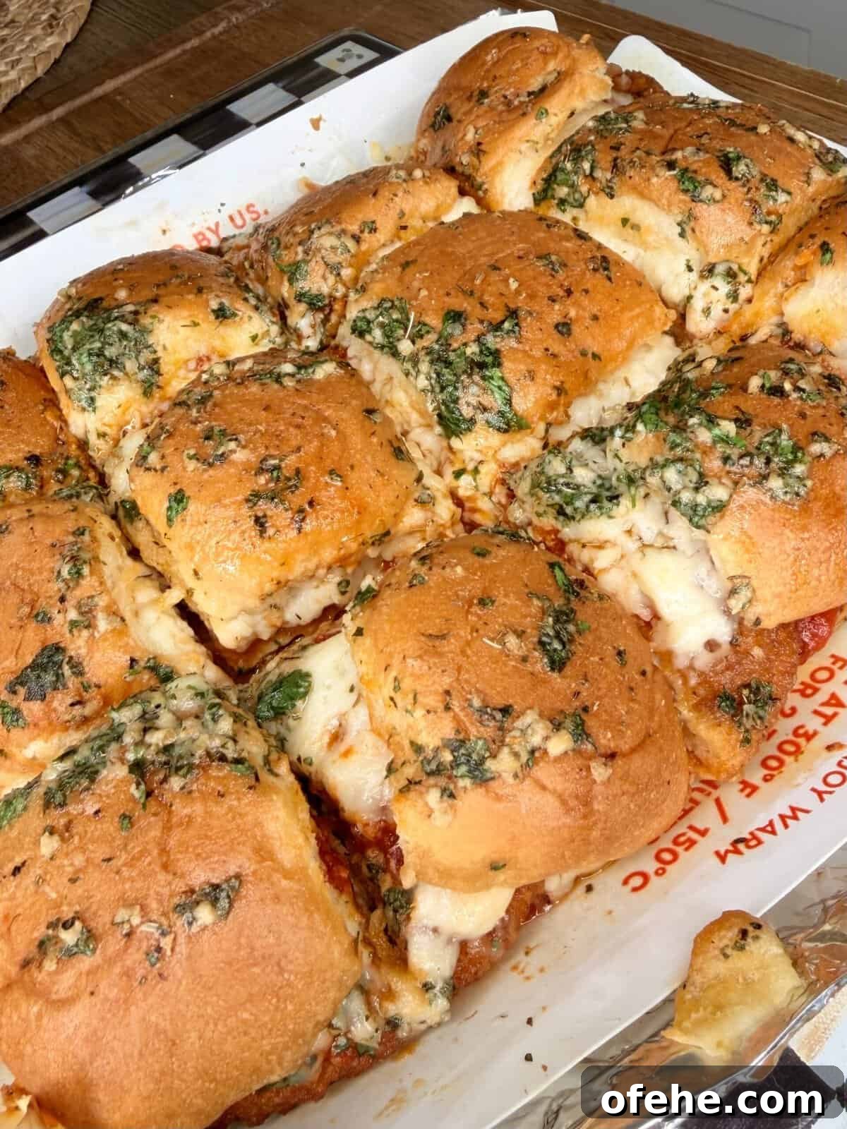 Golden, cheesy baked sliders topped with melted cheese, garlic, and herbs sit on a foil-lined tray. The sliders are closely packed, with gooey cheese oozing from between the soft, browned buns.