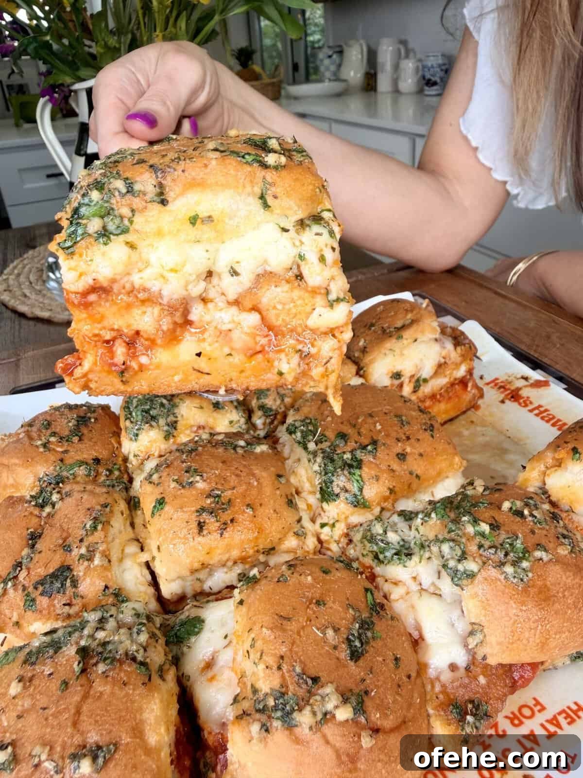 A hand lifts a cheesy, herb-topped sandwich from a tray of baked sliders filled with marinara sauce and melted cheese in a kitchen setting.