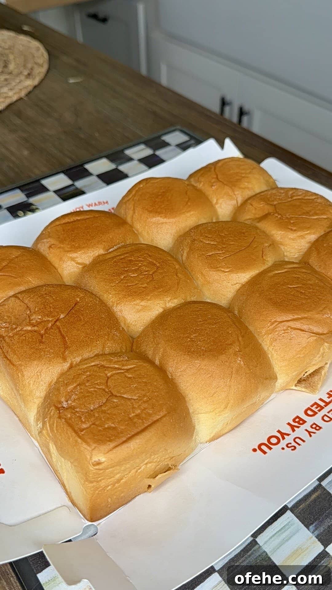 A batch of nine golden-brown dinner rolls sits on a white sheet of paper atop a checkered tray on a kitchen counter. The rolls look soft and freshly baked.
