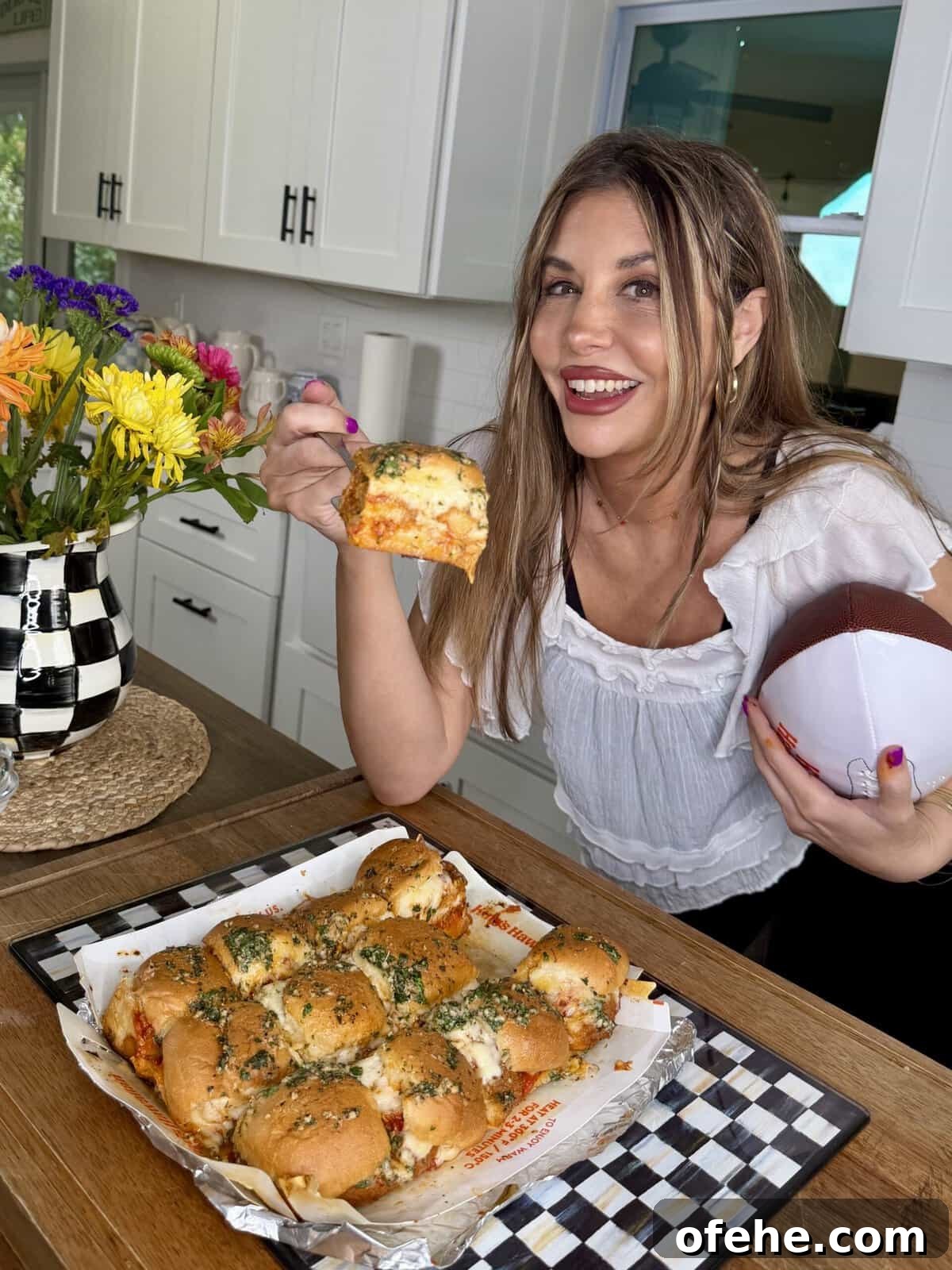A smiling woman holding a football in one hand and a cheesy garlic bread roll in the other, standing in a kitchen next to a tray of bread rolls and a vase of colorful flowers.