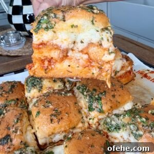 A hand lifts a cheesy, baked sandwich from a tray of pull-apart sliders, topped with herbs and melted cheese. The sandwiches are golden brown and layered with marinara sauce and mozzarella.