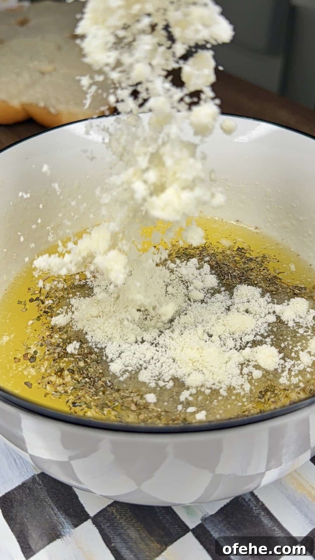 Grated cheese is being sprinkled into a white bowl filled with olive oil and herbs, likely for making a dip or dressing. A piece of bread is visible in the blurred background.