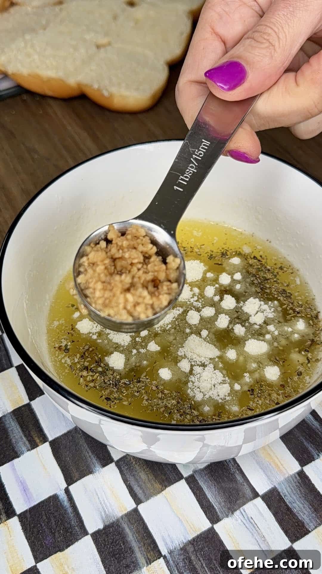 A hand with pink nail polish holds a tablespoon of minced garlic over a bowl containing melted butter, herbs, and grated cheese. Slices of bread are visible in the background.