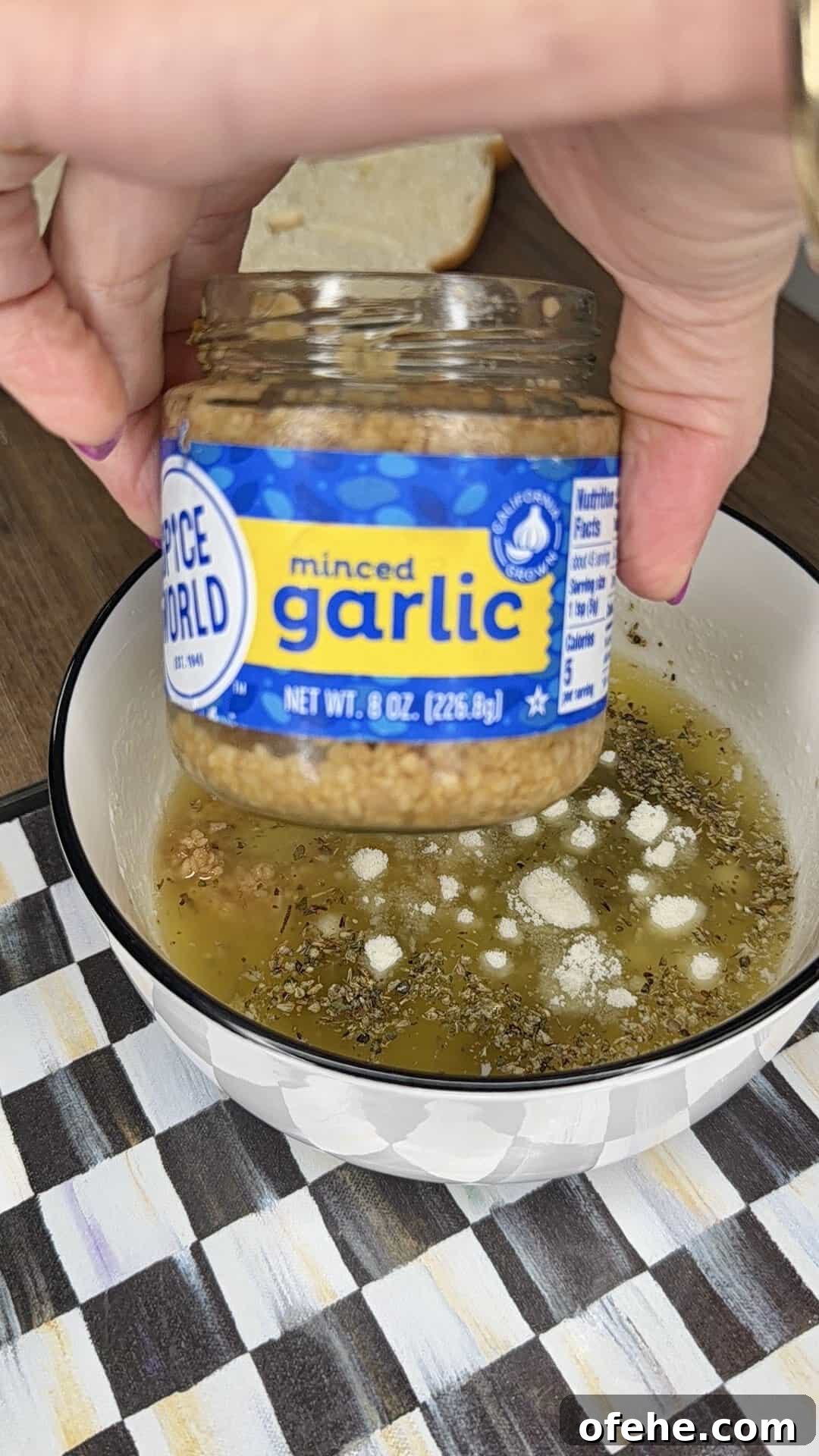 A hand holds a jar of minced garlic above a white bowl filled with olive oil, dried herbs, and seasonings on a checkered placemat. A slice of bread is visible in the background.
