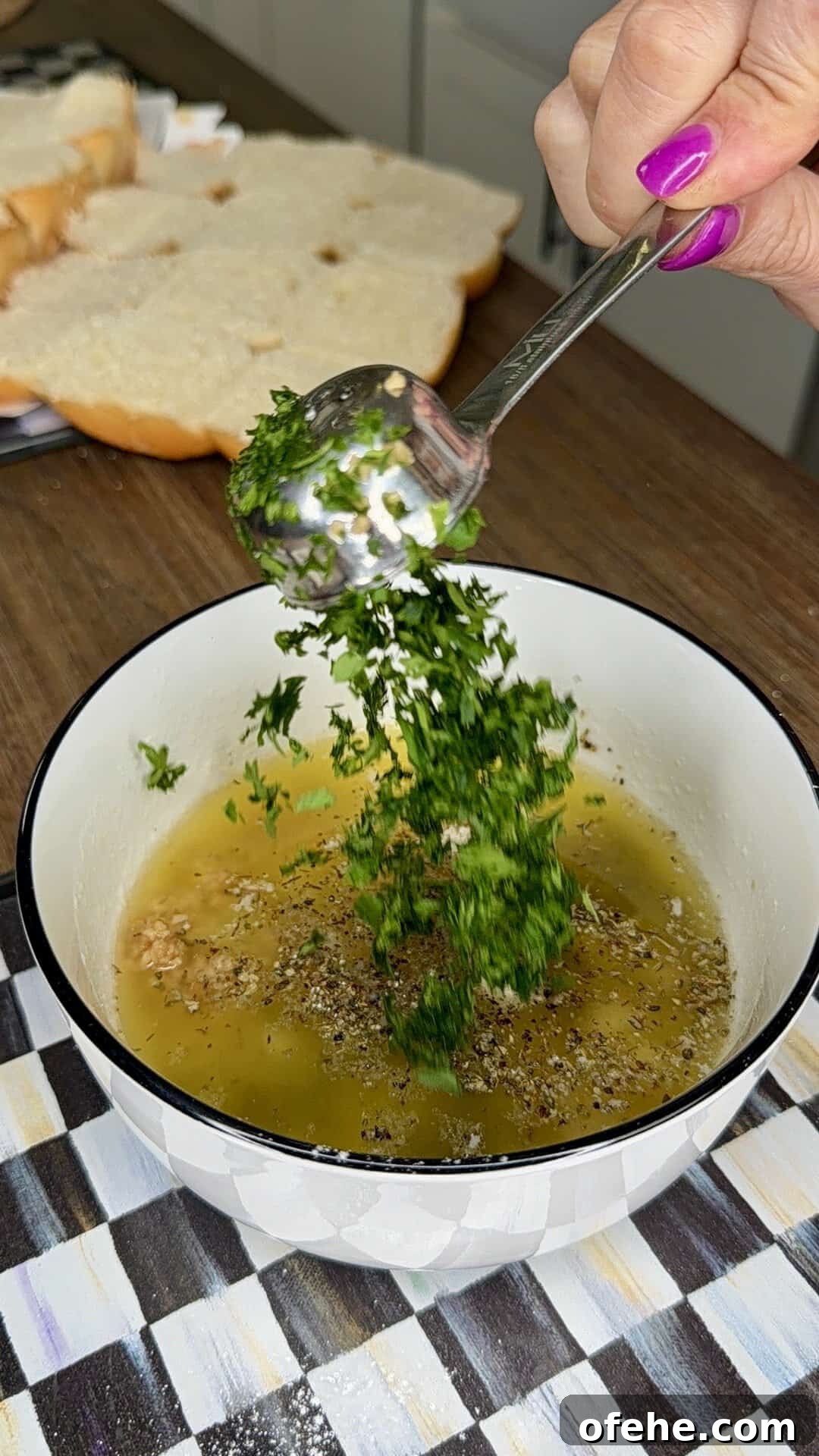A hand with pink nails adds chopped herbs from a spoon into a bowl of oil, pepper, and spices on a kitchen counter. Slices of bread are visible in the background.