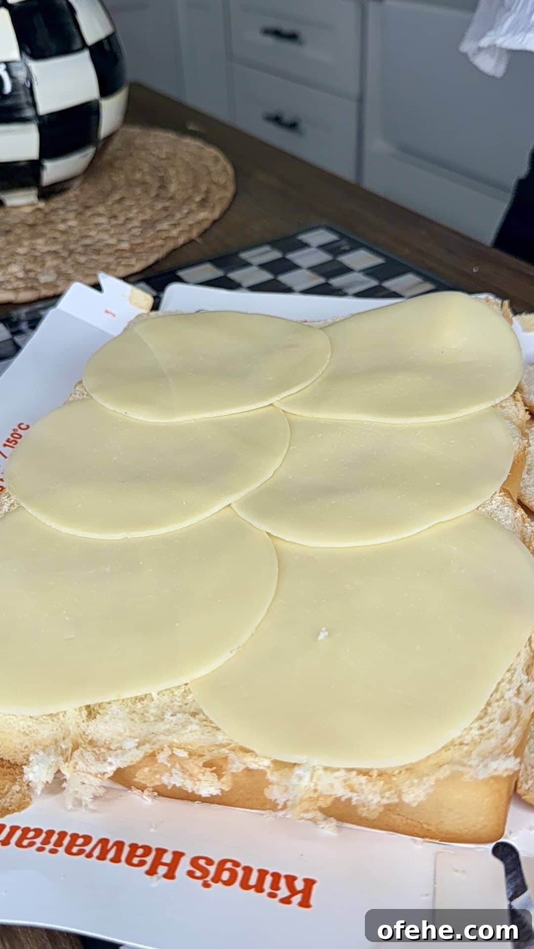 Six slices of provolone cheese are laid out on a piece of white bread covered with mayonnaise, on top of Kings Hawaiian bread packaging, with a kitchen counter and checkered decor in the background.