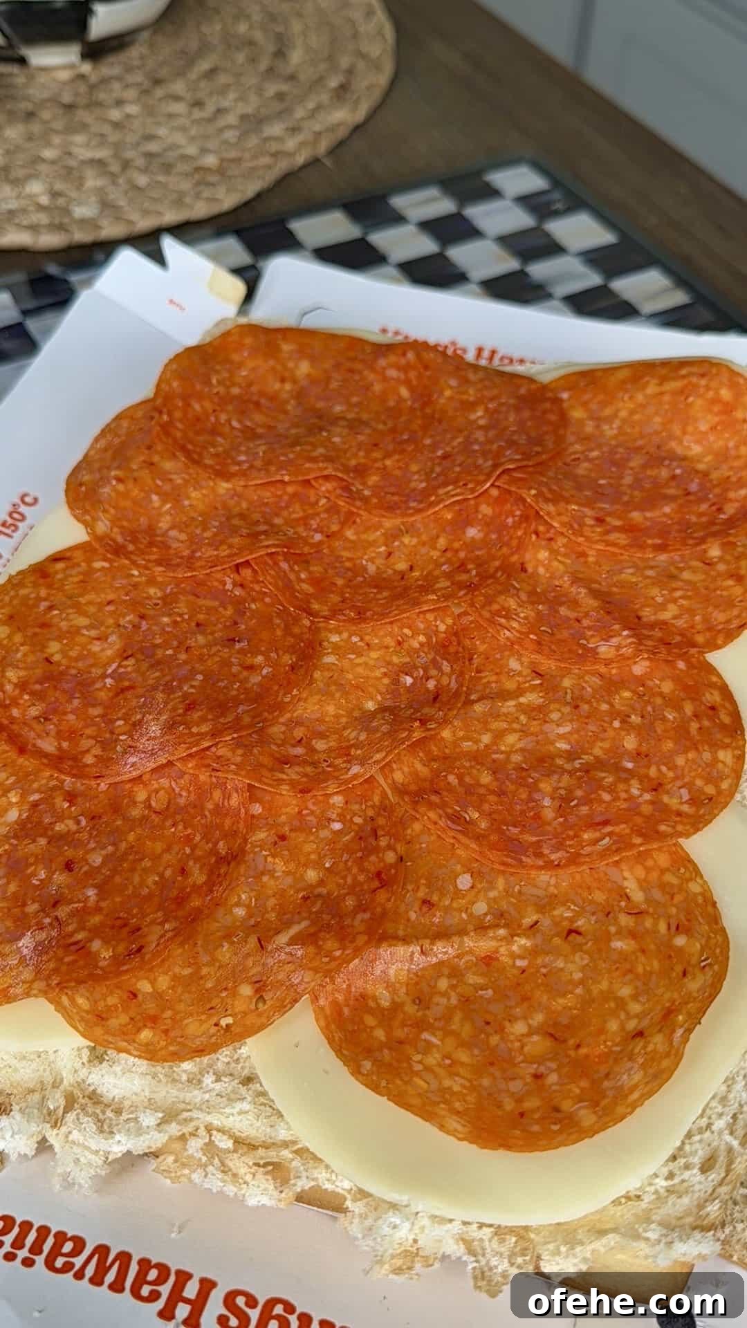 Several slices of pepperoni are layered over white cheese on a sandwich roll, sitting on deli paper, with part of a woven placemat and checkered surface visible in the background.