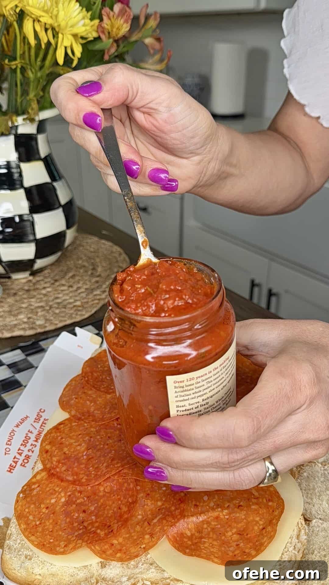 A person with purple nail polish spreads red sauce from a jar onto a sandwich with cheese and pepperoni slices. A vase of flowers and checkered pitcher are in the background on a kitchen counter.