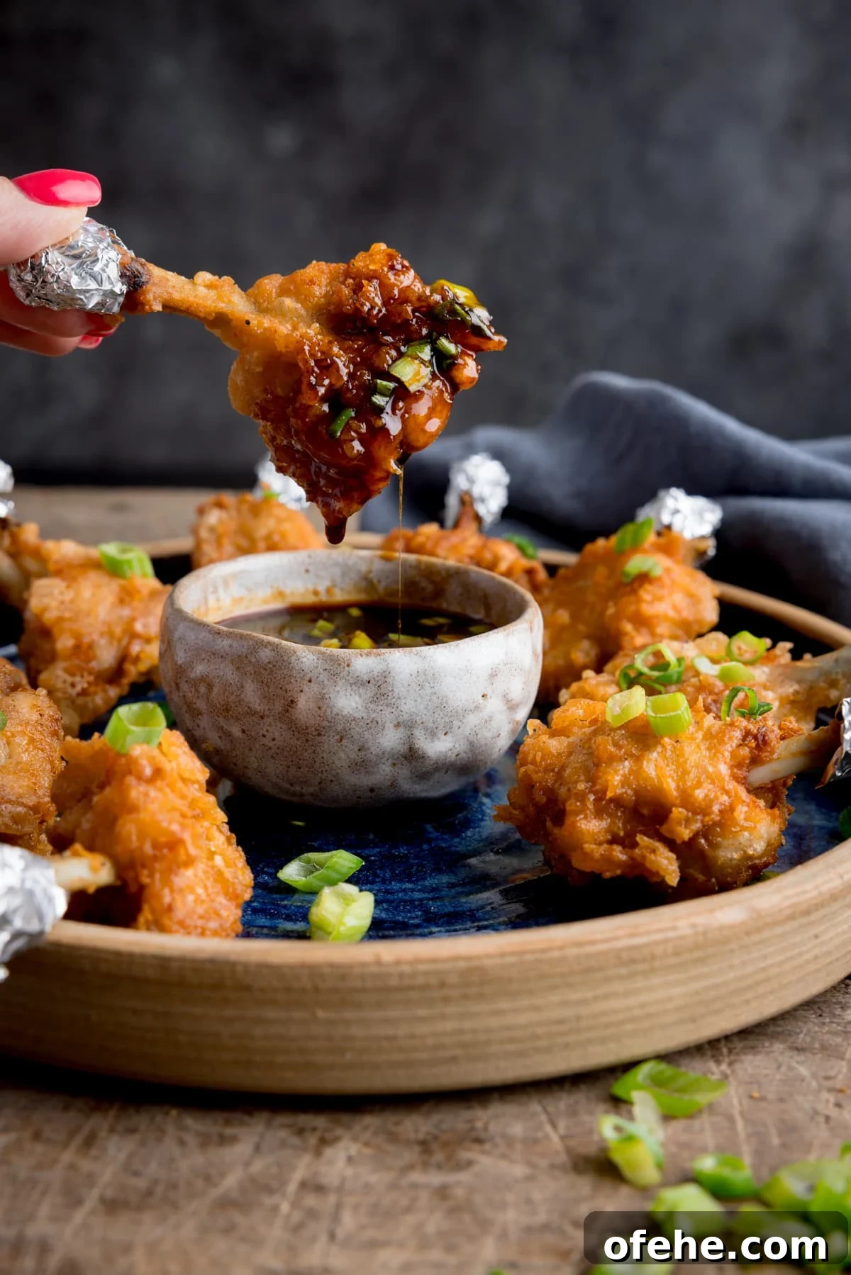 Crispy Chicken Lollipops Recipe: Finger-Licking Party Snack 2 A plate of chicken lollipops with a bowl of spicy dipping sauce. One of the wings is being dipped into the sauce and is dripping. The wing is being held by fingers with pink-painted fingernails.